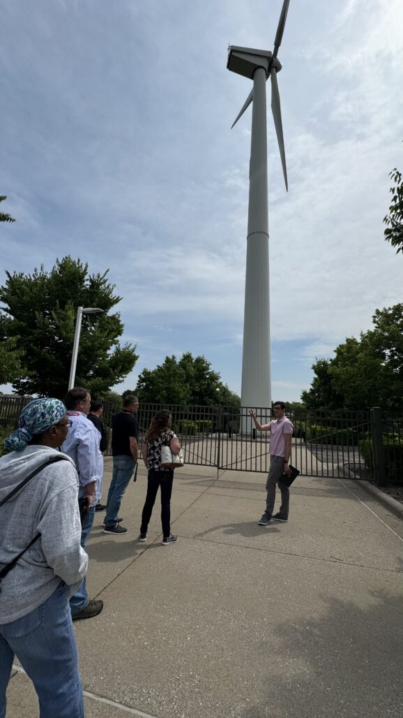 Group listening to a male engineer explain parts of a wind turbine in front of a large wind turbine. 