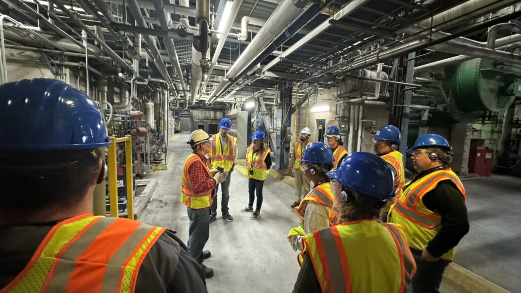 group in hard hats and safety vests in a large energy plant room with many pipes